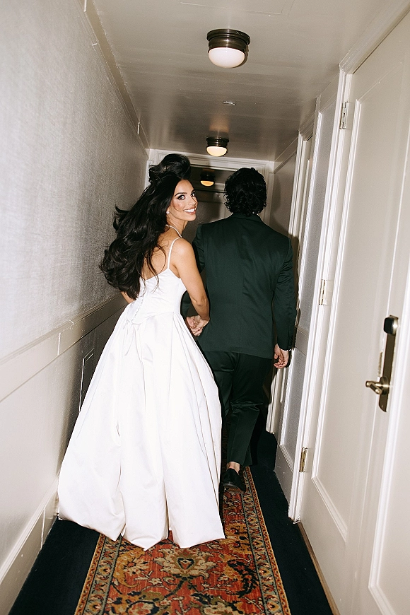 Couple portrait of bride and groom walking away holding hands in a hotel hallway, bride looking back with gown train under ceiling lights