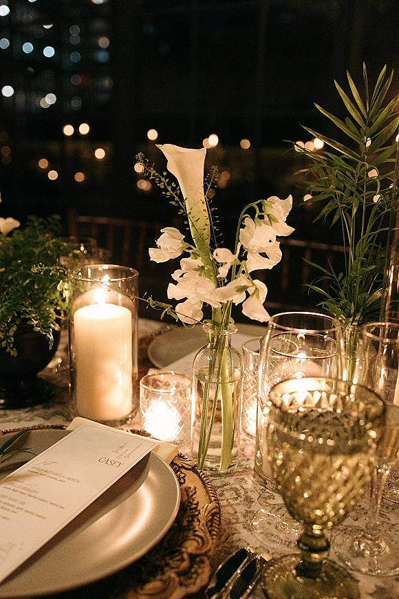 Reception tablescape with wedding table centerpiece of white flowers and greenery, pillar and votive candles on lace tablecloth under string lights at dusk