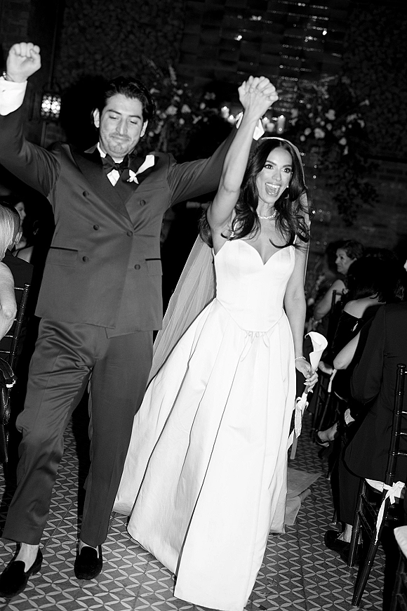Wedding recessional as bride in strapless gown and cathedral veil and groom in tux raise hands, walking past cheering guests indoors