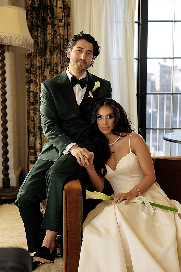 Couple portrait of bride and groom holding hands, seated on a sofa by window light with city skyline; bride wears a satin dress