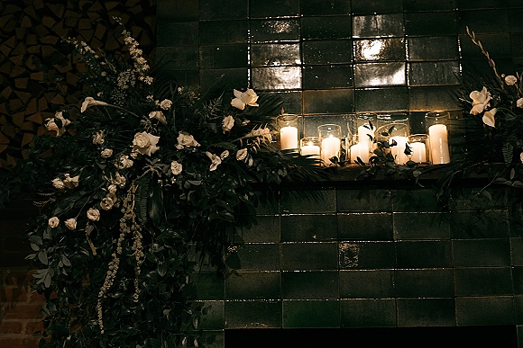 Wedding candle decor with glass hurricane candles and pillar clusters, white roses and greenery garland against a dark green tile wall