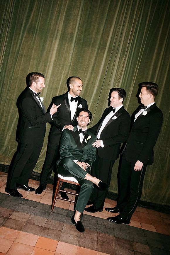 Groomsmen portrait of a groom and groomsmen in black tuxedos and bow ties, groom in green double-breasted suit before a green velvet curtain