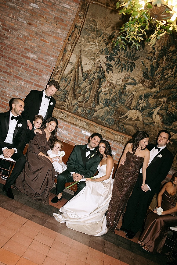 Wedding party portrait with bride and groom seated on a bench, surrounded by tuxedos and brown dresses against a brick wall backdrop