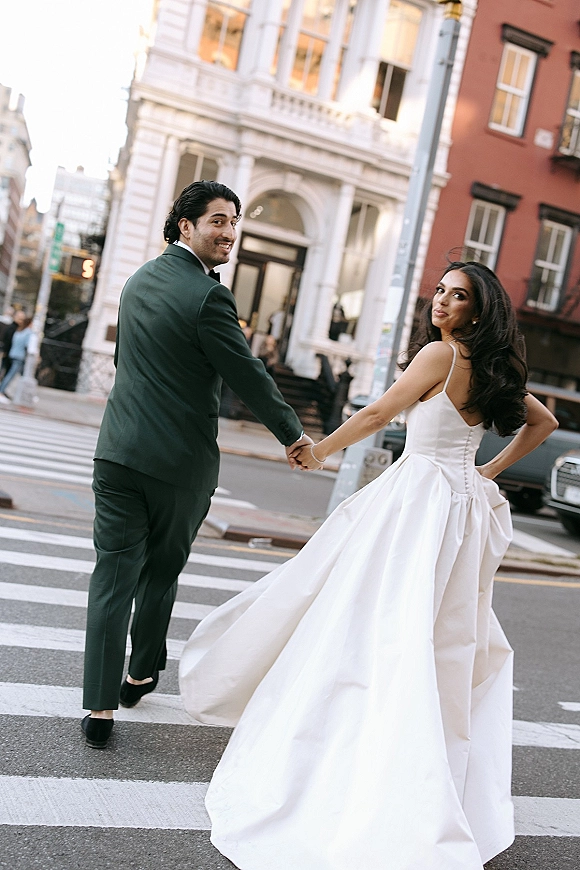 Couple portrait of bride and groom holding hands on a city crosswalk, bride in ball gown and groom in black tuxedo looking back
