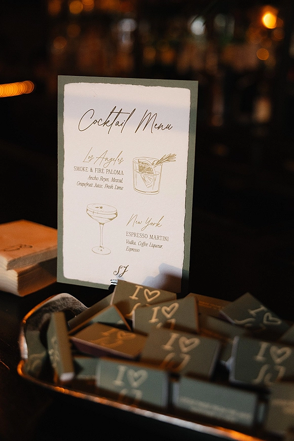 Wedding cocktail menu card with calligraphy text and illustrated drinks, clipped on a tray beside favor boxes in dim bar bokeh lighting