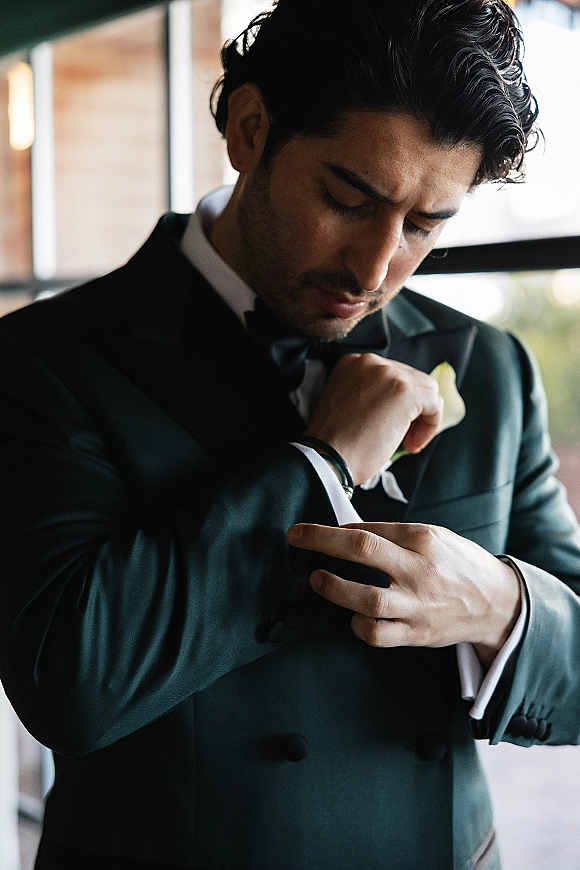 Groom portrait in window light, groom getting ready as he adjusts cufflinks on a dark green tuxedo with black bow tie and boutonniere