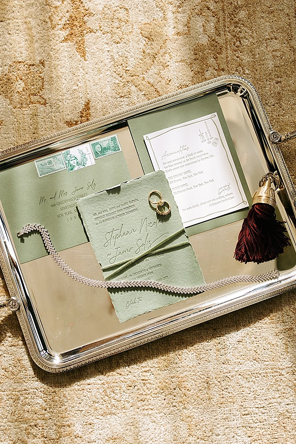 Wedding invitation suite with green wedding invitations, RSVP card, envelopes, wax seal, ribbon and rings on a silver tray over patterned carpet