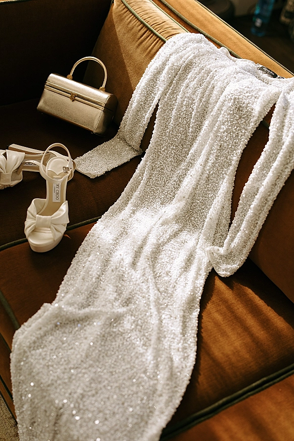 Wedding dress and beaded wedding dress draped on a brown velvet sofa with white bow heels and a gold clutch in a living room interior
