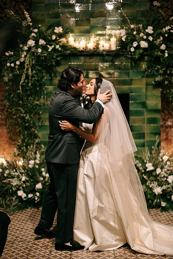 Wedding kiss during an indoor ceremony, bride in satin dress and veil with groom in suit under greenery arch, candlelit tiled wall backdrop
