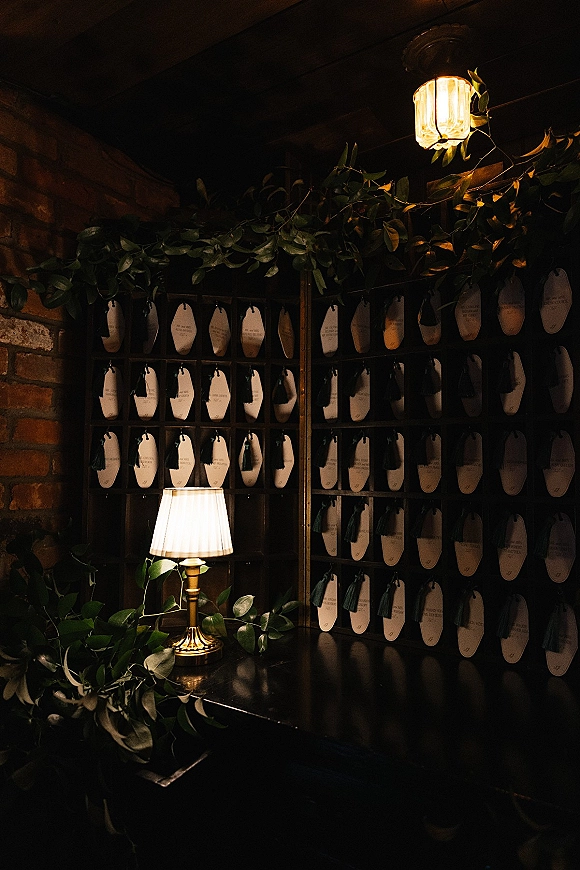 Wedding escort card display with an escort card wall of tassel name cards in a wood cubby, greenery garland, and warm light against brick wall