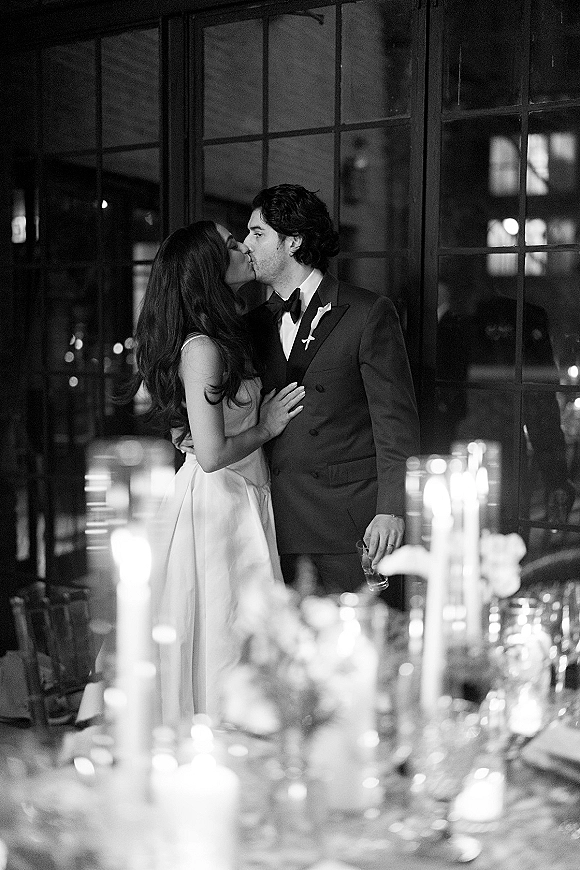 Wedding kiss portrait in a black and white wedding photo, bride in strapless gown kissing groom in tuxedo by windows with candlelit table behind