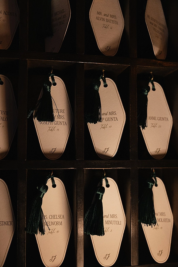 Wedding escort cards with vintage hotel key style tags and black tassels, calligraphy names hung on hooks in a dark wood cubby display