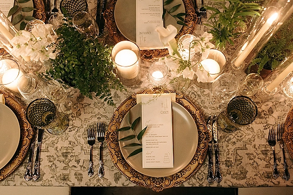 Reception tablescape with wedding place setting, ornate gold chargers on patterned tablecloth, green goblets, menus, place cards, and taper candles