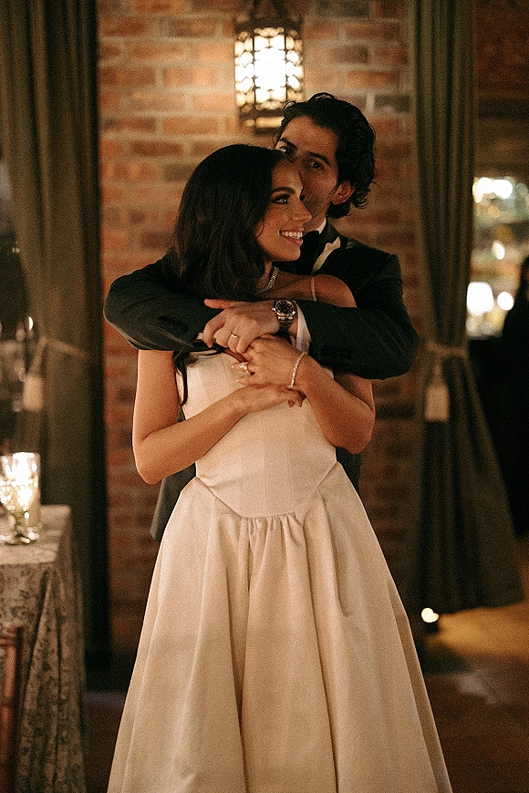 Couple portrait of bride and groom embrace, her strapless gown and rings visible, by brick wall with lantern and candlelit reception tables