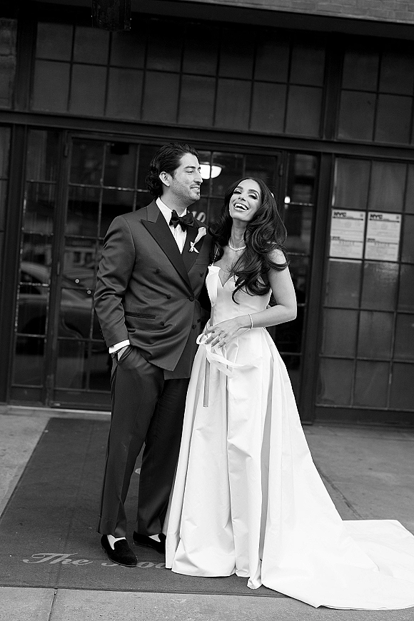 Couple portrait in a black and white wedding portrait, bride in strapless gown and veil laughing with groom in tuxedo by tall windows