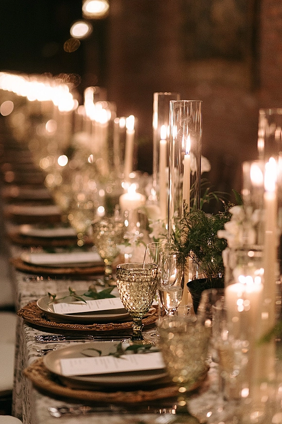 Reception tablescape with candlelit wedding reception glow, taper candles in glass hurricanes, white florals and greenery garland in a dim room with bokeh lights