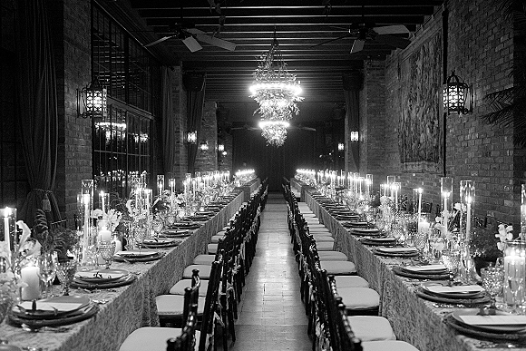 Reception tablescape with long banquet tables set in candlelight, taper candles and floral greenery centerpieces under chandeliers in a brick-walled room
