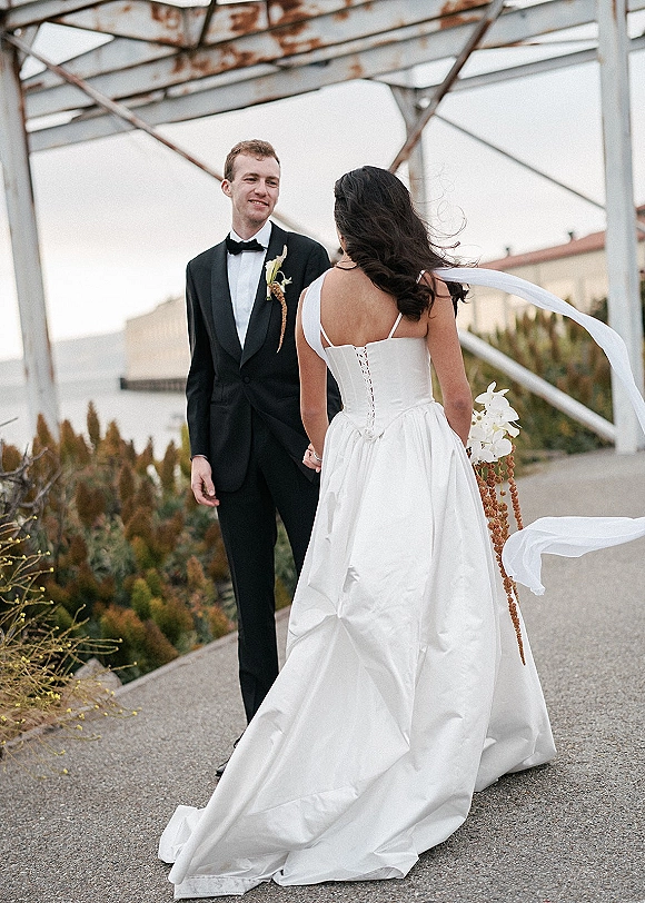 Couple portrait of bride and groom holding hands, her veil blowing as she approaches him in an industrial outdoor walkway under overcast sky