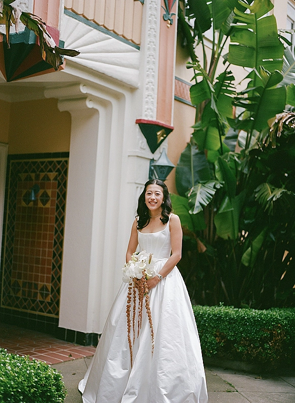 Bridal portrait of a smiling bride holding bouquet, wearing a white satin gown and necklace beside tropical plants and an archway