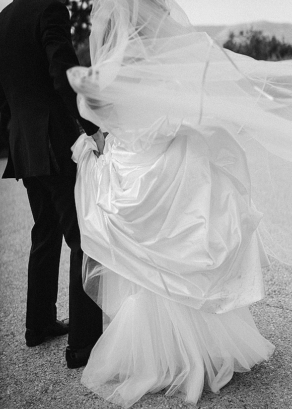 Wedding couple portrait in black and white, bride and groom embracing as her veil blows over them on a gravel path by trees