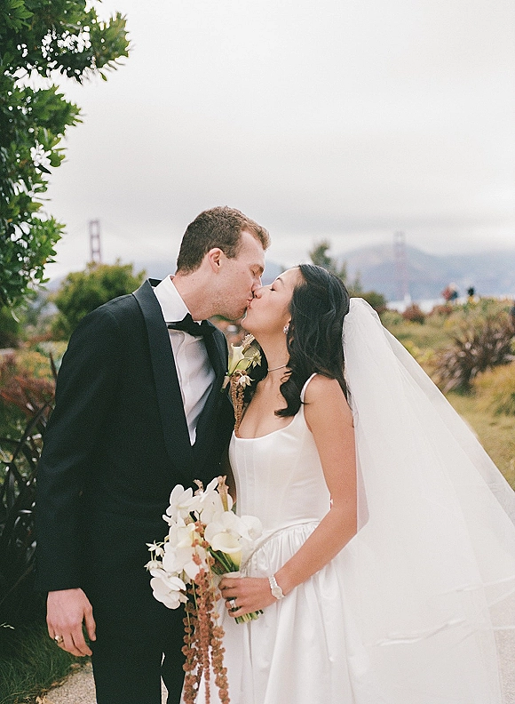 Wedding kiss as bride and groom embrace, her long veil flowing and orchid bouquet in hand against greenery and cloudy sky backdrop
