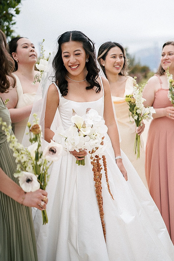 Bride portrait holding bouquet, smiling in a veil and wedding dress with bridesmaids in pastel dresses under open sky and trees