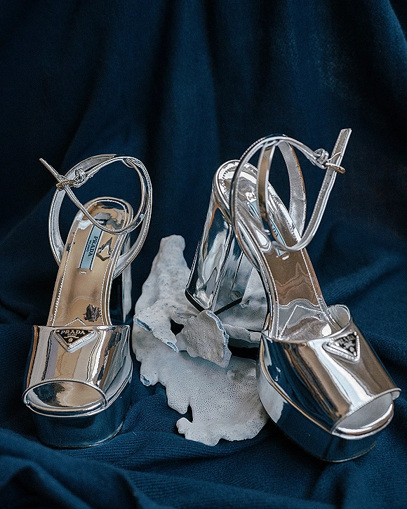 Bridal shoes, silver wedding heels with ankle straps and designer logo plaque on coral rock prop against blue fabric drape