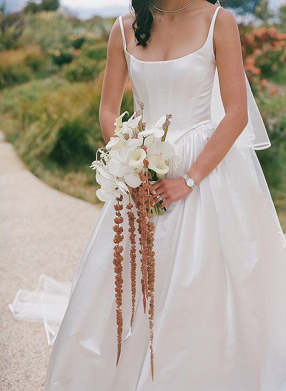 Bridal gown in simple satin with spaghetti straps, bride holding cascading calla lily and orchid bouquet on a gravel garden path