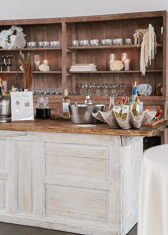 Wedding bar setup with a bar menu sign, wine and liquor bottles, glassware, and candles on wooden shelves in an indoor reception space