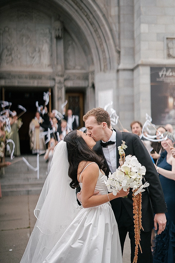 Wedding kiss as newlyweds share a kiss on church stone steps, bride in veil holding cascading orchid bouquet as guests cheer outside