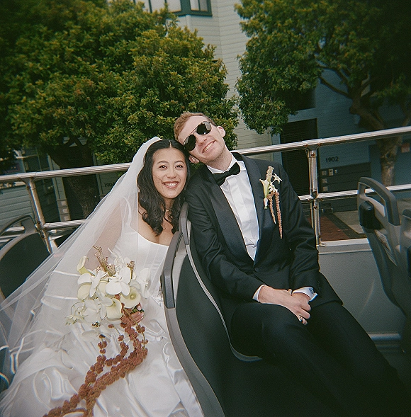 Couple portrait of bride and groom seated on a wedding getaway bus seat, wearing sunglasses; she holds a cascading calla lily bouquet by trees.
