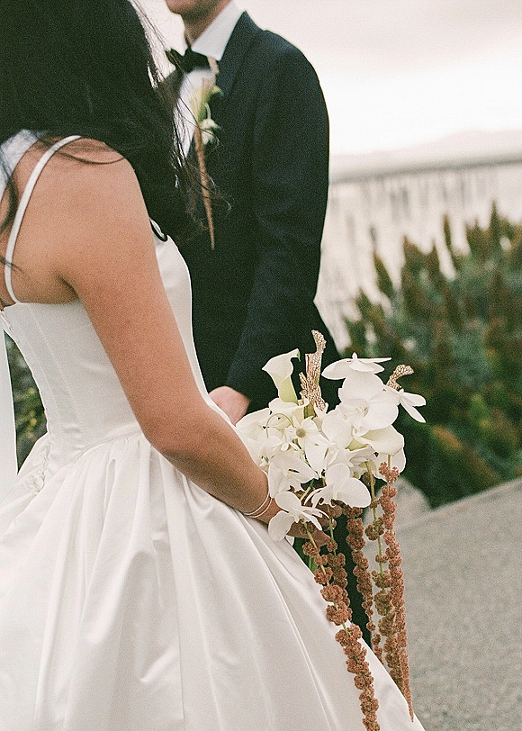 Wedding couple portrait with bride holding bouquet of white orchids and calla lilies, standing on a stone path by the ocean coastline