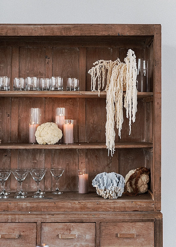 Reception bar decor with wedding bar styling on wood shelves, featuring martini and rocks glasses, cylinder candles, and dried hanging florals