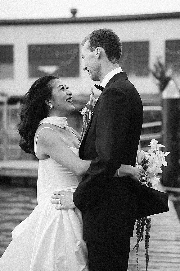 Couple portrait in black and white wedding portrait style, bride in strapless dress embraces groom with bouquet on a patio with string lights