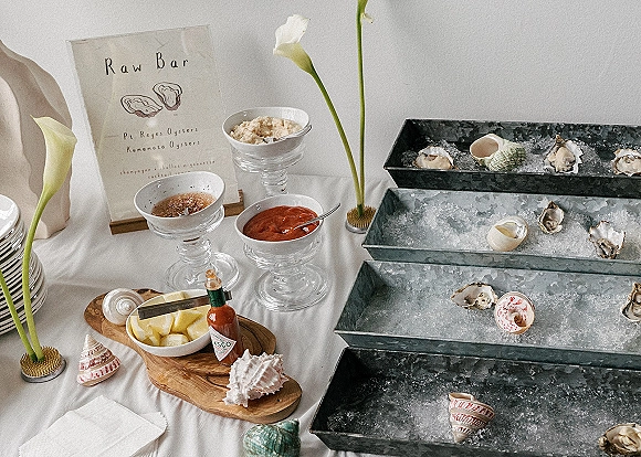 Wedding raw bar with oysters on ice in galvanized trays, martini glass sauces and lemon wedges beside a menu sign on white linens