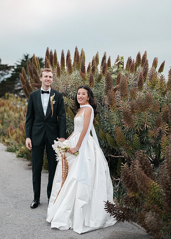 Couple portrait of bride and groom holding hands, bride in strapless satin gown with veil and orchid bouquet on a garden path