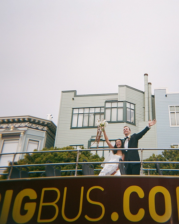 Newlywed portrait, just married photo of bride lifting bouquet with ribbons as groom waves from an open-top bus beside row houses