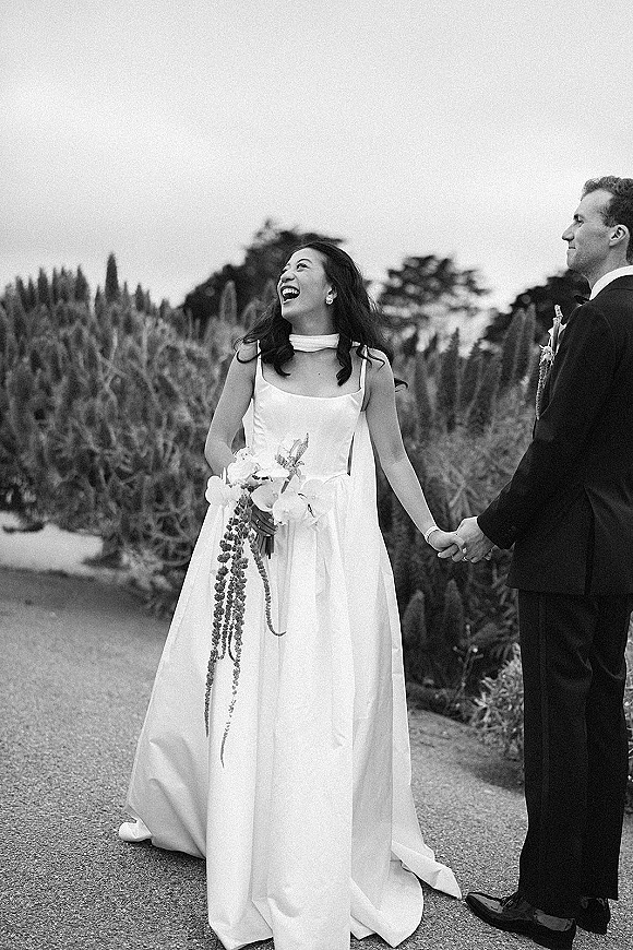 Couple portrait in a black and white wedding portrait, bride laughing while holding hands with groom, bouquet and suit in a garden walkway