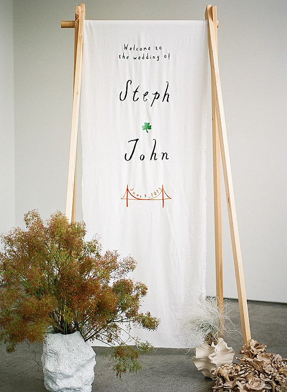 Wedding welcome sign on a wooden stand with calligraphy lettering and a green shamrock motif, with dried florals in a ceramic vase on floor