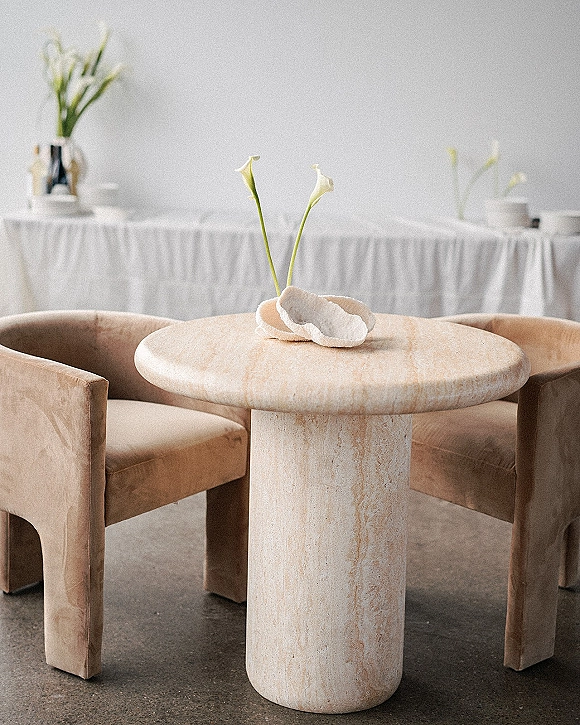 Reception lounge seating with upholstered armchairs around a stone pedestal table, accented by calla lily bud vase beside a white draped buffet table