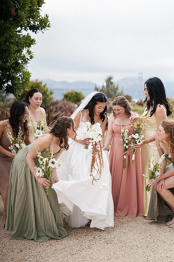 Bridesmaids portrait of bride with bridesmaids holding bouquets around her veil, mismatched dresses on a garden gravel path under cloudy sky