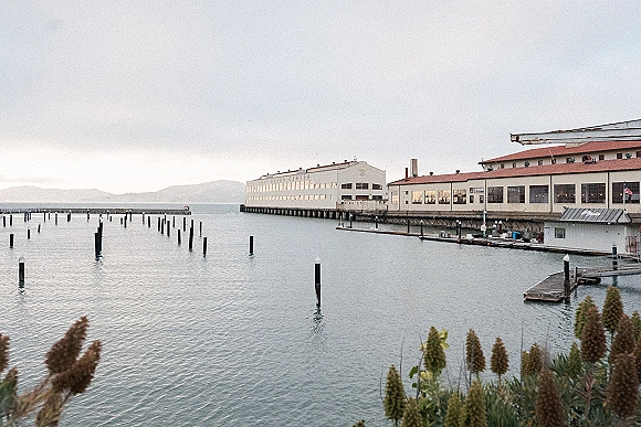 Waterfront venue exterior at a wedding venue waterfront with dock pier pilings and an industrial building, calm bay water and distant hills beyond