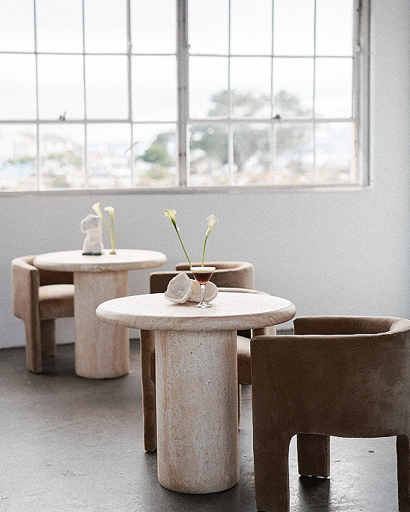 Cocktail lounge seating with round stone tables, calla lily bud vases and a cocktail glass beside folded napkins in an industrial loft venue