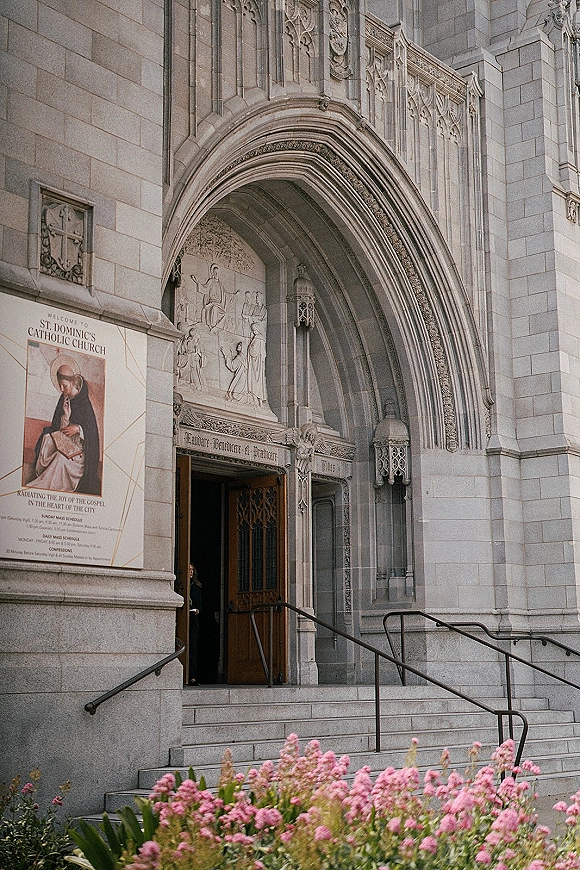 Church entrance with a stone archway and wooden doors, carved relief above steps with handrails and pink flowers by the facade