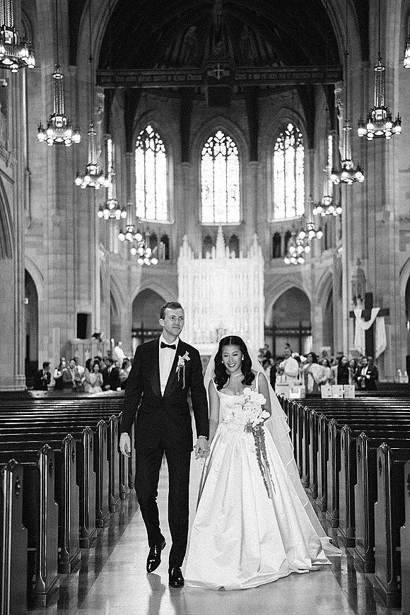 Wedding recessional as bride and groom walk the church aisle hand in hand, her veil and bouquet flowing past pews and stained glass