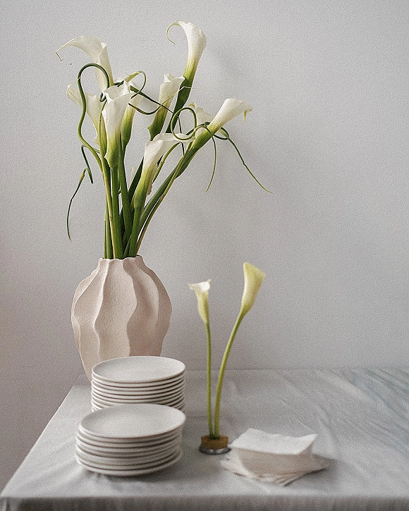 Reception tablescape with calla lily centerpiece in a white ceramic vase, paired with bud vases, stacked plates, and crisp linens on a tabletop