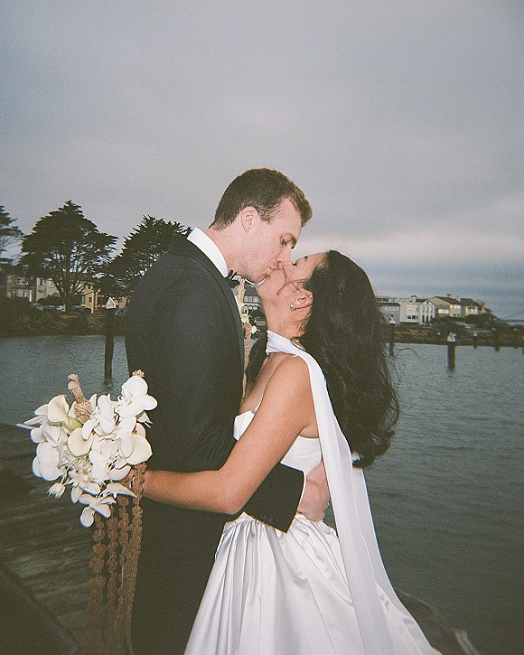 Wedding kiss portrait of bride and groom kissing on a wooden pier, bride holding cascading white orchid bouquet, waterfront under cloudy sky