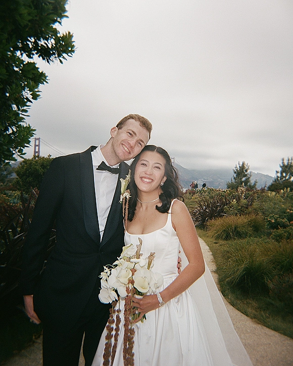 Couple portrait of bride and groom smiling and hugging on a garden path, bride holding a cascading calla lily bouquet in satin dress