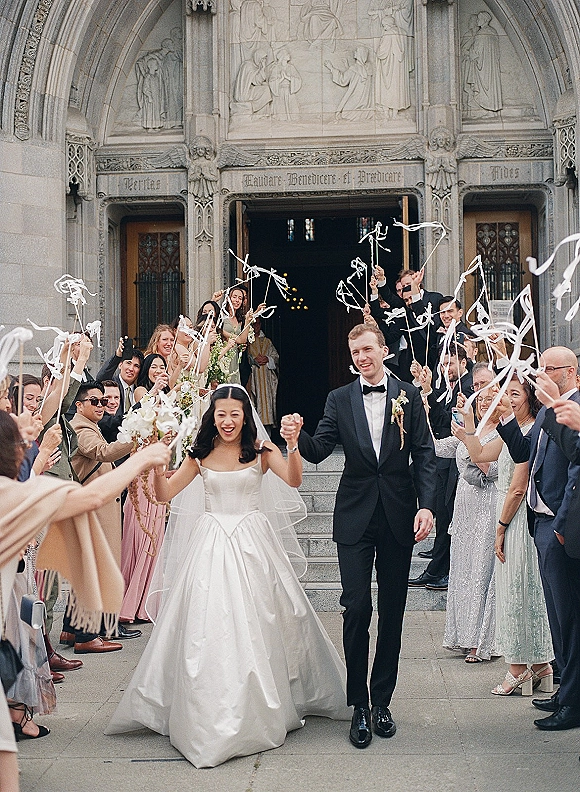 Wedding recessional as bride and groom exit the church, guests waving ribbon wands on stone steps, bride holding bouquet and veil flowing