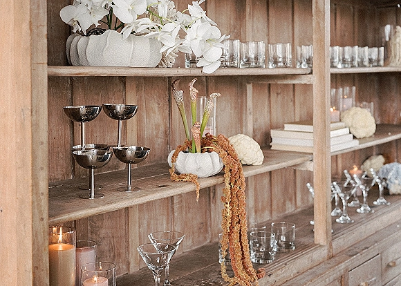 Wedding bar decor on rustic wood shelves with mixed glassware, votive candles, white orchids, calla lilies, and pumpkins against a wood wall